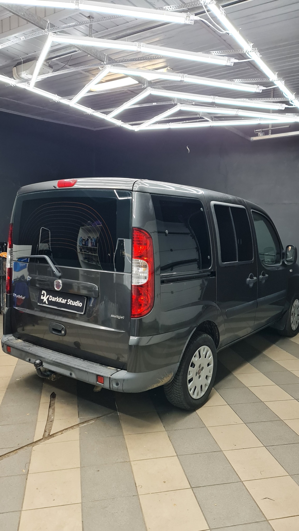 Szary samochód typu van marki Fiat Doblo z przyciemnionymi szybami, zaparkowany w studiu detailingowym DarkKar Studio, widok z tyłu, oświetlony lampami LED na suficie.