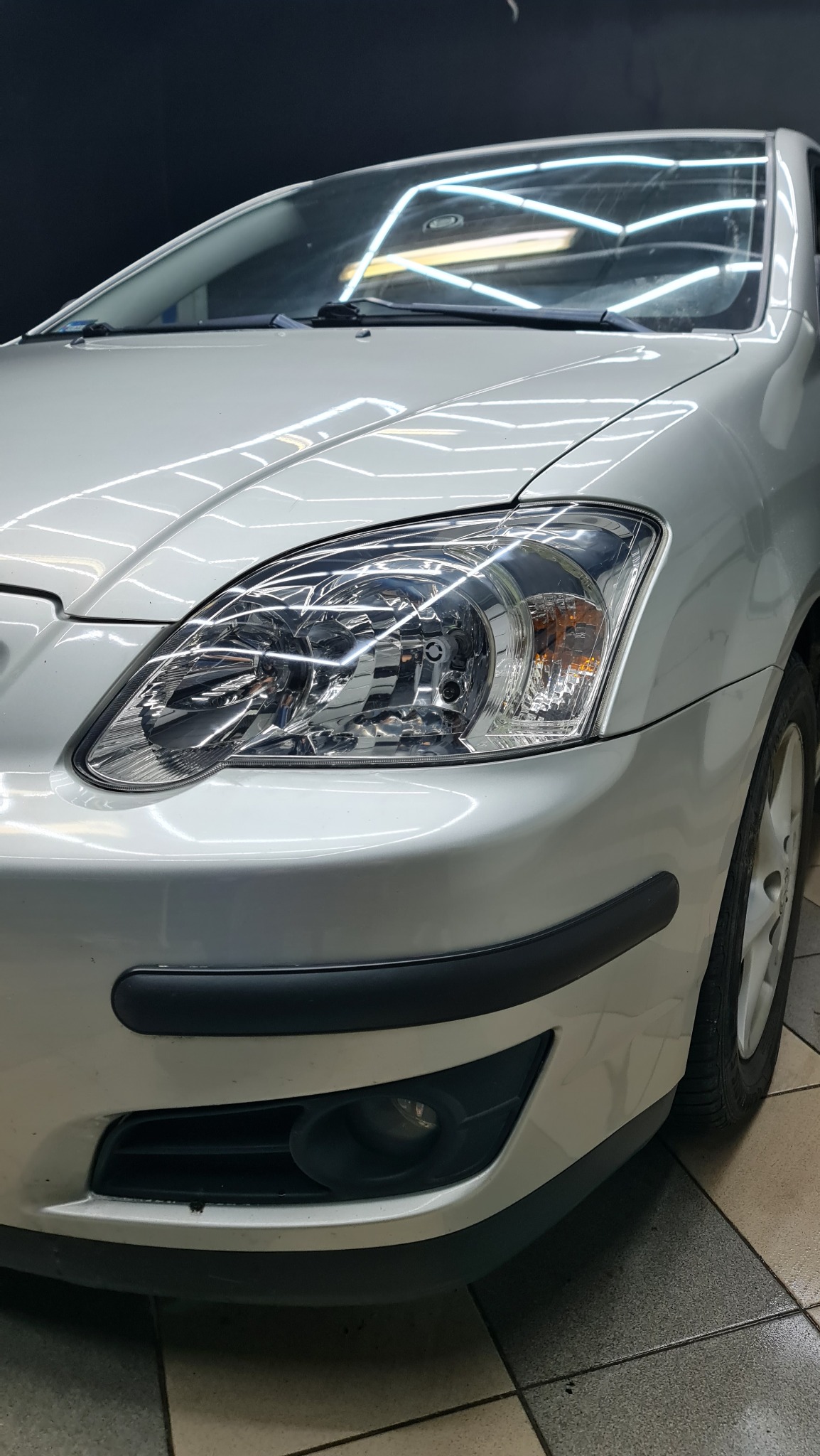 Srebrny samochód, widok z bliska na przedni reflektor i fragment błotnika z czarną listwą ochronną, widoczne odbicia światła na karoserii.