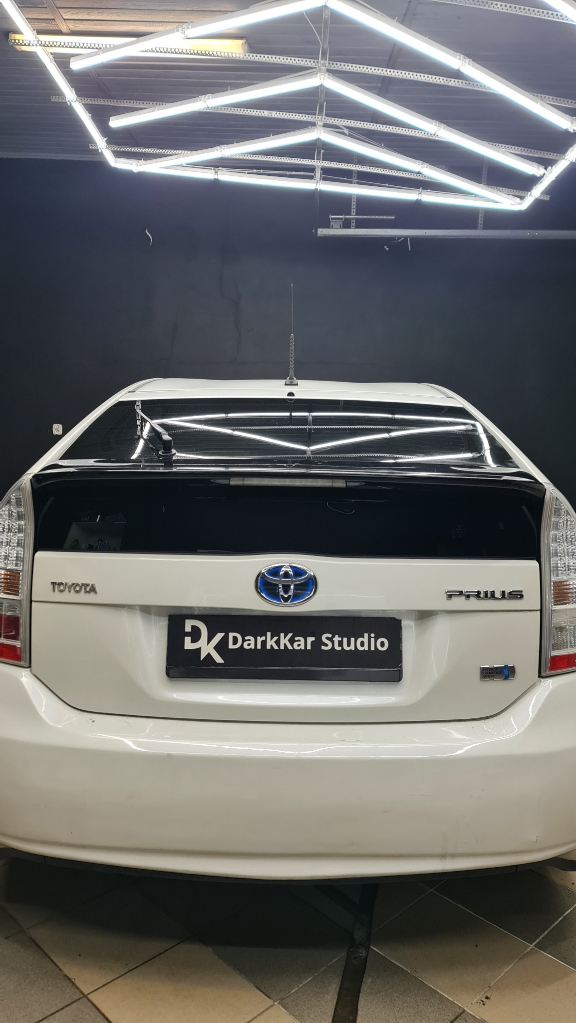 Biała Toyota Prius z przyciemnioną tylną szybą, widoczny znak firmy DarkKar Studio, oświetlenie sufitowe w tle.