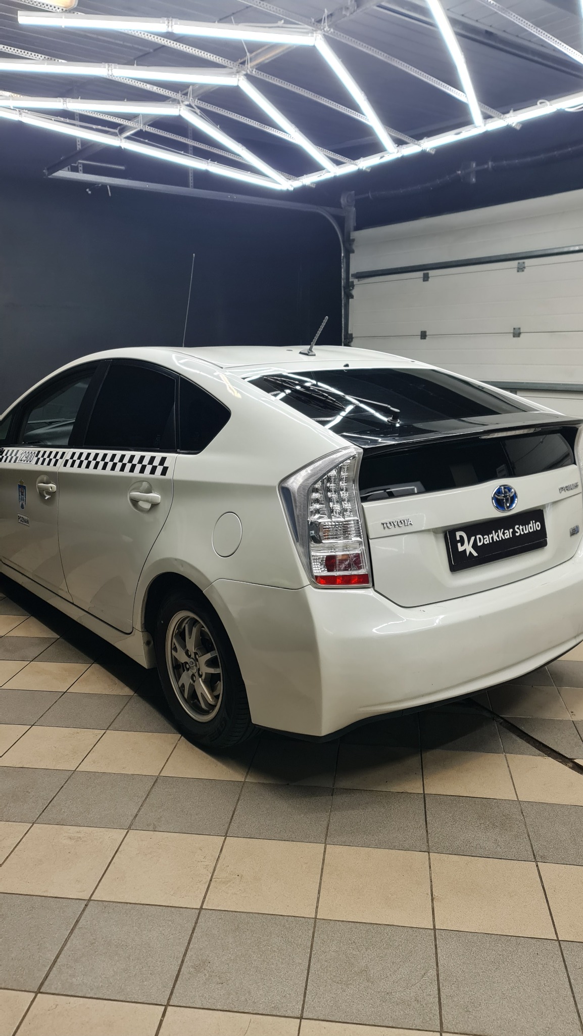 Biała Toyota Prius z folią w szachownicę na tylnej szybie, zaparkowana w dobrze oświetlonym garażu z logo 'DarkKar Studio' na klapie bagażnika.