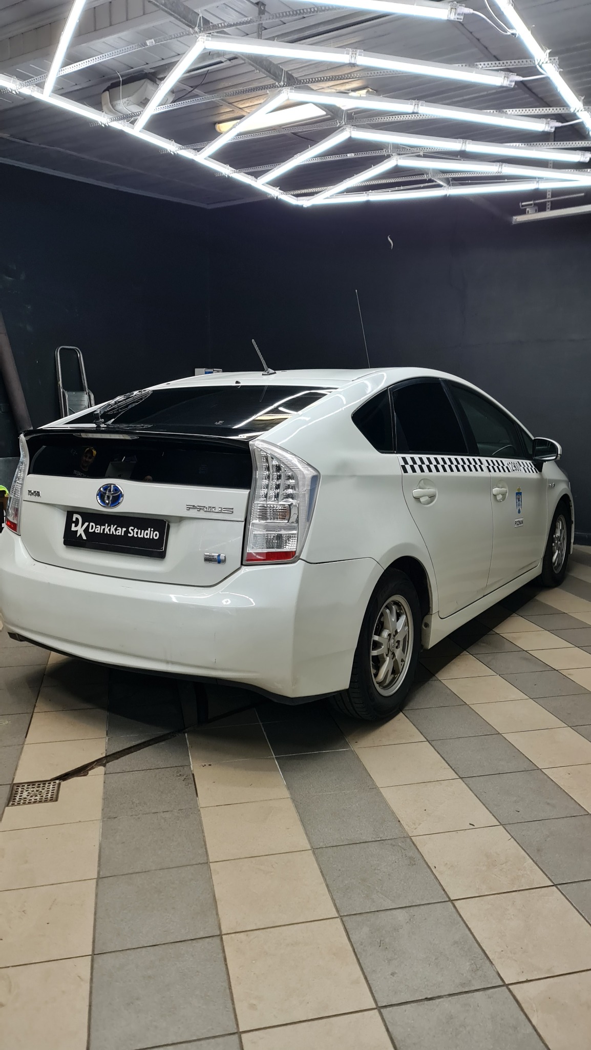 Biały samochód Toyota Prius z czarnym spoilerem i przyciemnionymi szybami, oklejony w czarno-białą szachownicę, stojący w dobrze oświetlonym garażu z czarną ścianą w tle.