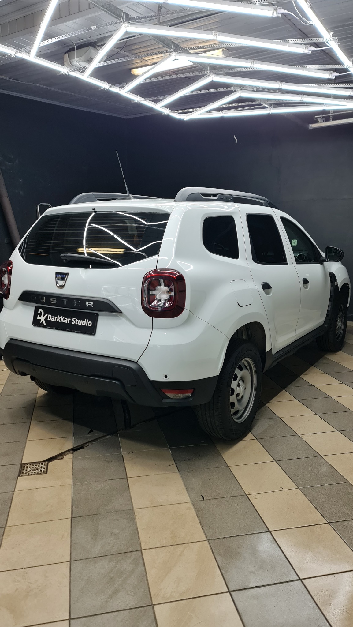 Biały samochód Dacia Duster widziany z tyłu w dobrze oświetlonym garażu z logo DarkKar Studio na klapie bagażnika.