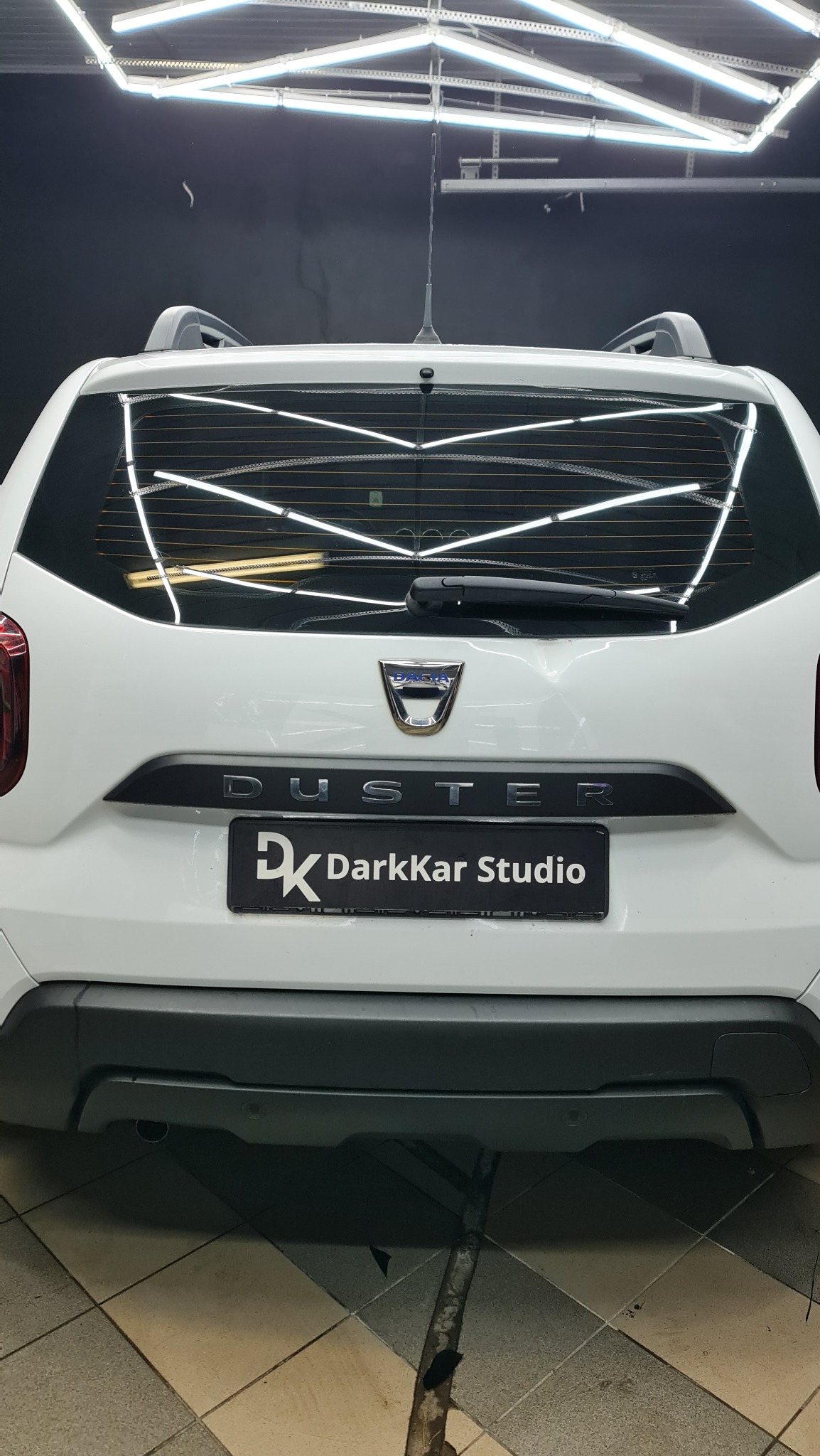 Tył białego samochodu Dacia Duster z widocznym logo firmy DarkKar Studio na tablicy rejestracyjnej, przyciemnione szyby w górnej części, oświetlenie warsztatowe odbija się na szybie.