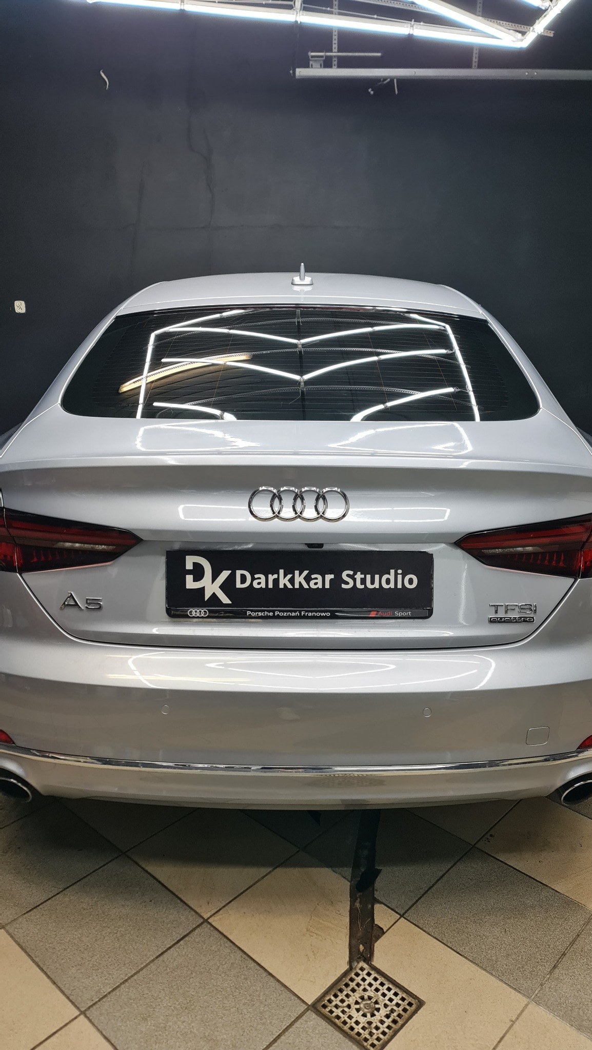 Srebrne Audi A5 z widocznym logotypem DarkKar Studio na tylnej klapie, oświetlenie studyjne odbija się w szybie, widoczna kratka ściekowa na podłodze.