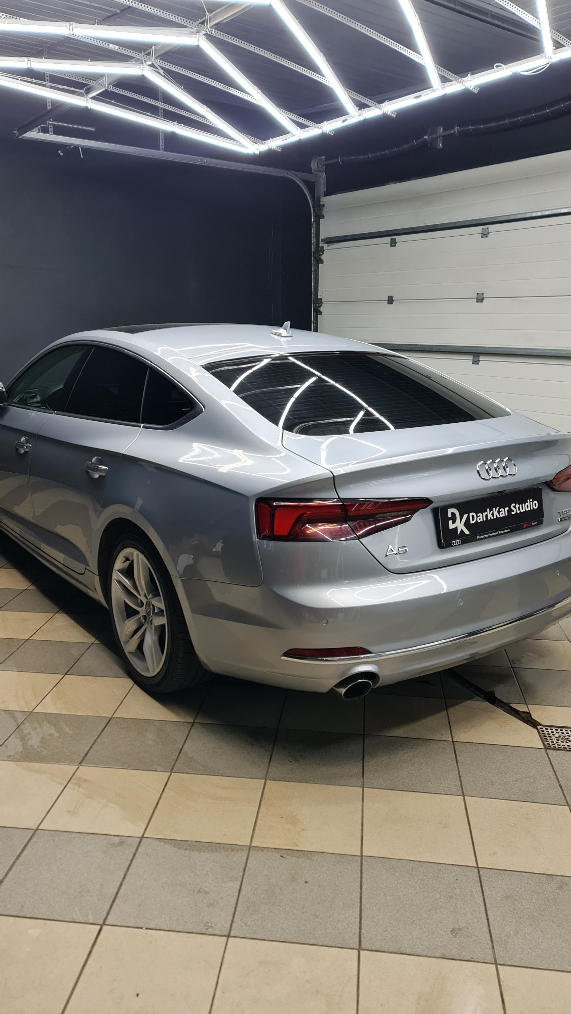 Srebrny samochód Audi A5 po zabiegu przyciemniania tylnych szyb, widoczny w garażu z jasnym oświetleniem LED na suficie.