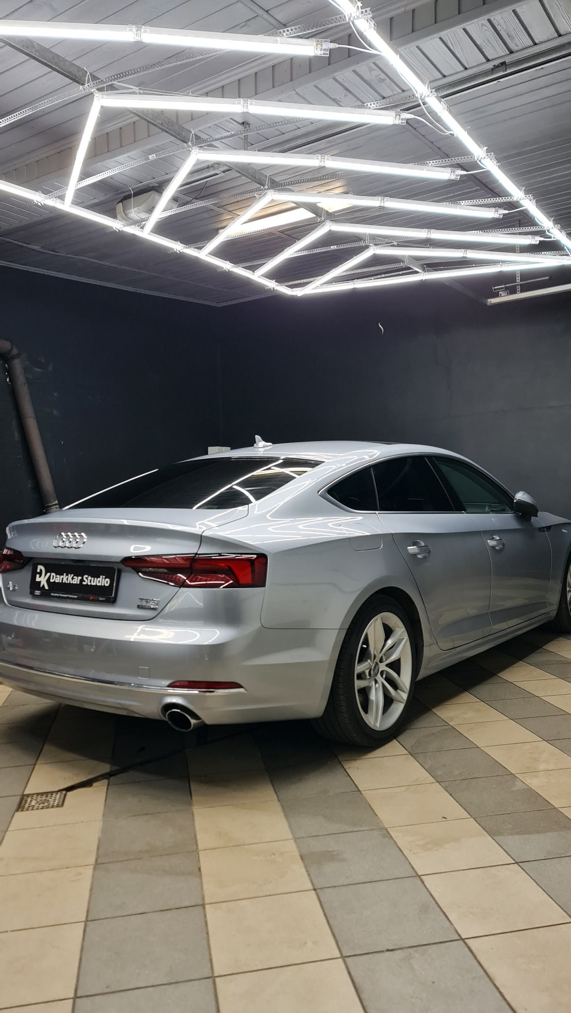 Srebrne Audi A5 po zabiegu przyciemniania szyb, widoczne z tyłu, w dobrze oświetlonym garażu z kratkowaną podłogą i neonowym oświetleniem sufitowym w geometryczne wzory.