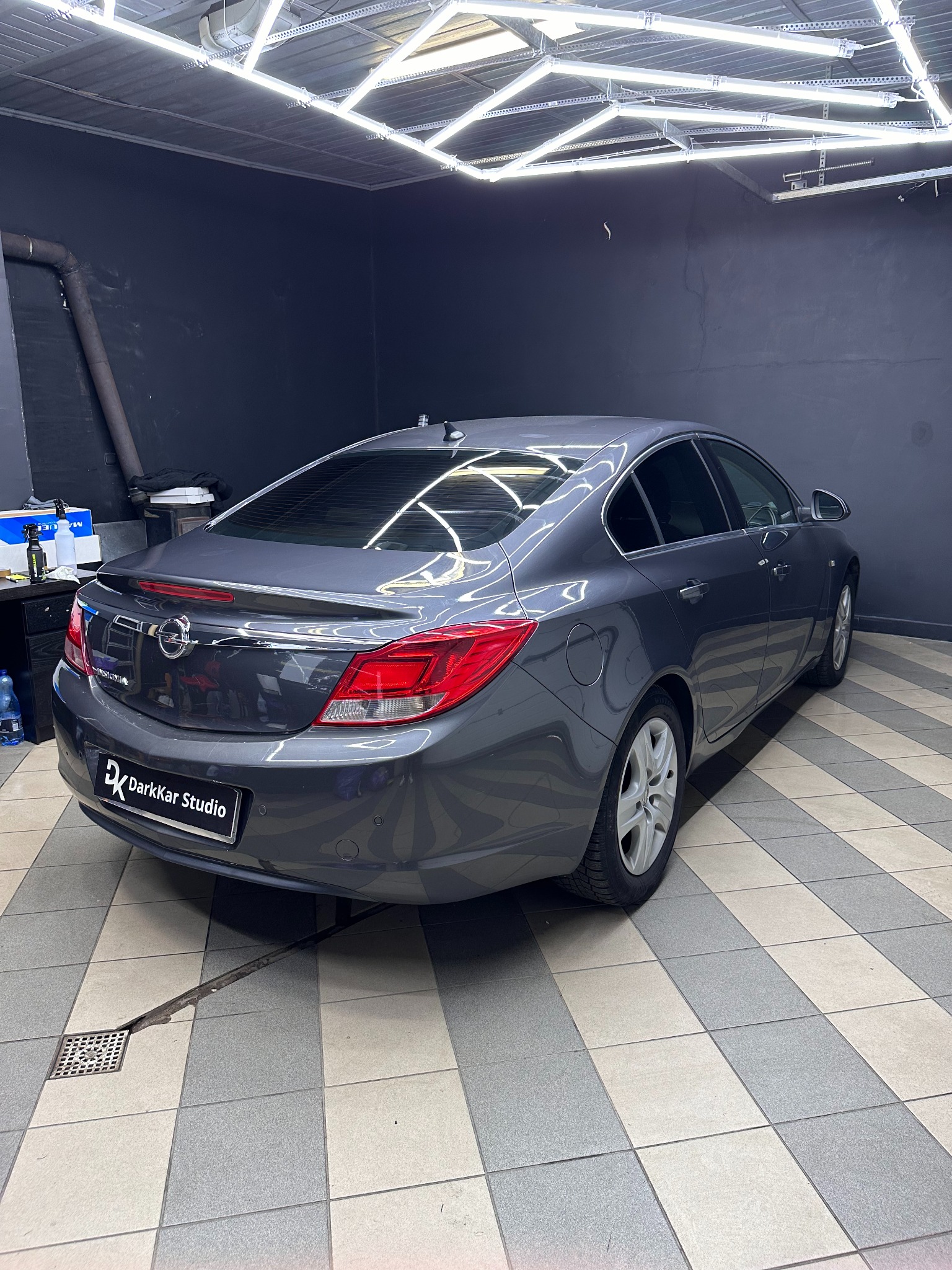Szary Opel Insignia po przyciemnieniu szyb, widoczny z tyłu w dobrze oświetlonym garażu z kratką ściekową w podłodze i logo 'DarkKar Studio' na tablicy.
