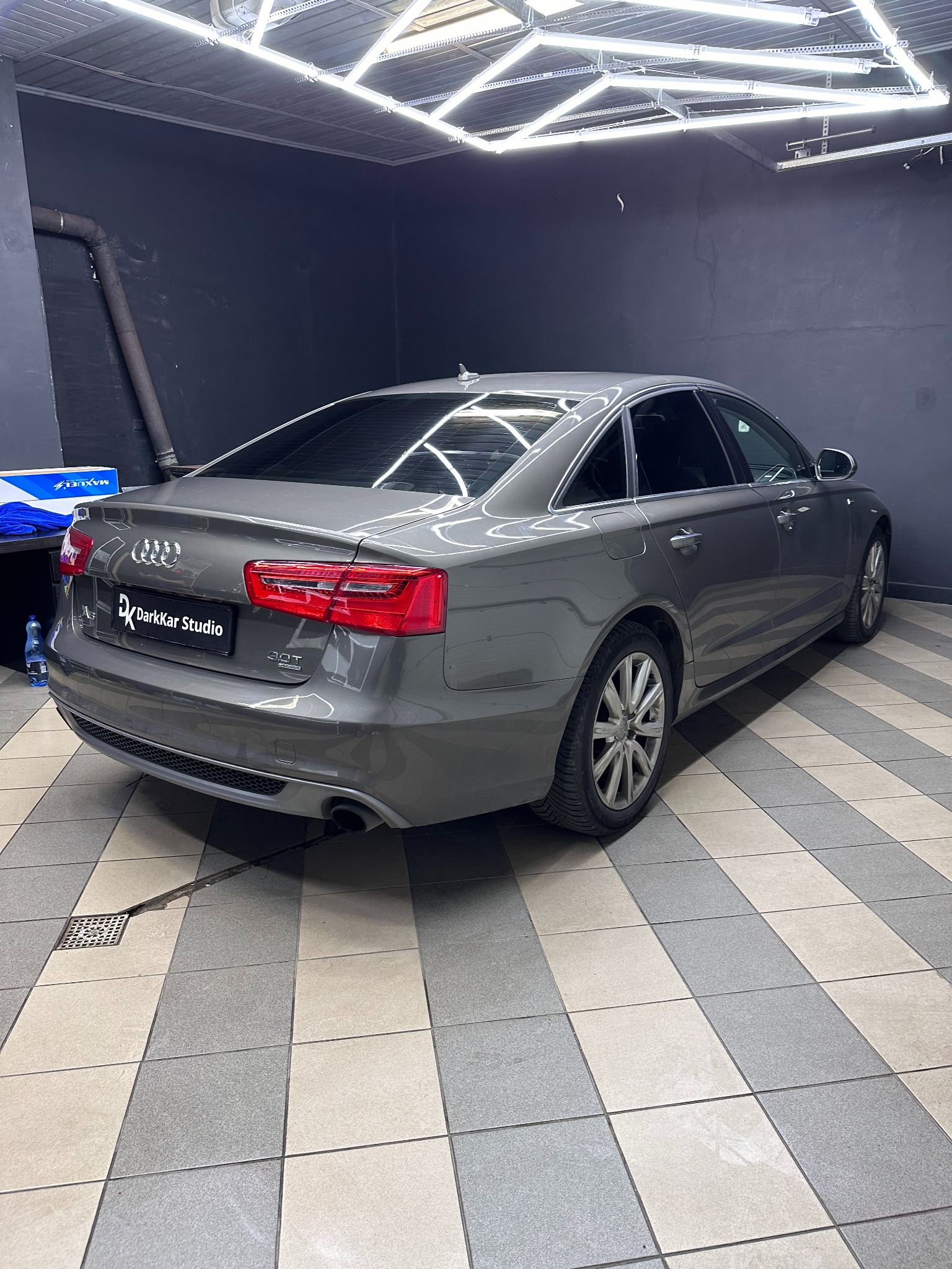 Szary Audi A6 po zabiegu przyciemniania szyb, widoczne ciemne szyby tylne i boczne, zaparkowane w studiu detailingowym DarkKar Studio z charakterystycznym oświetleniem sufitowym.
