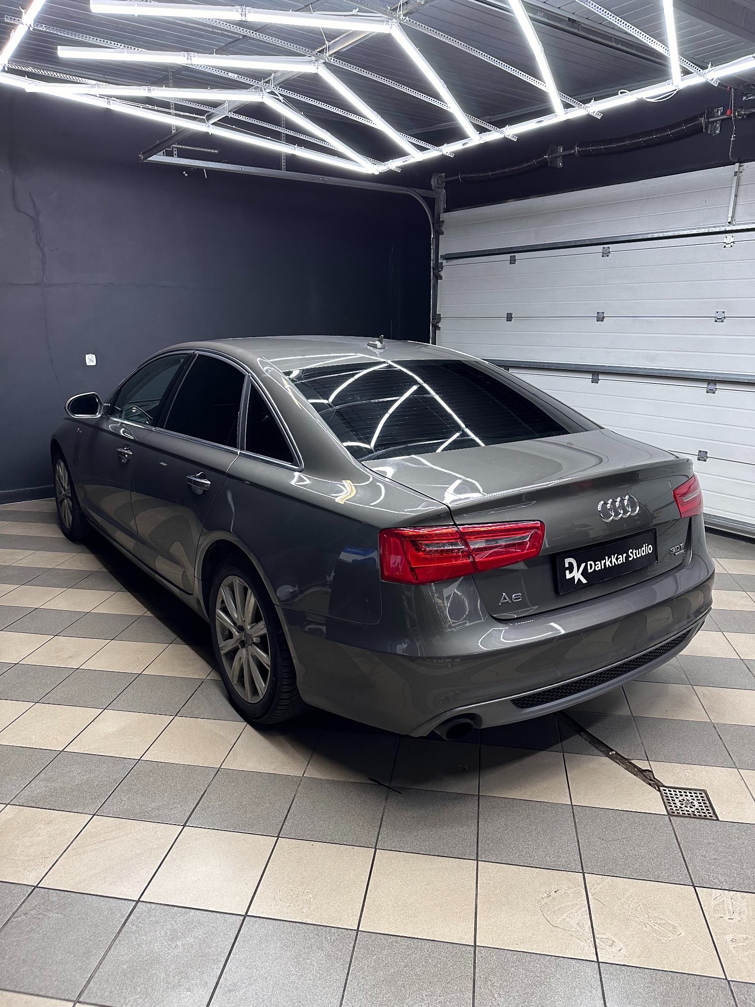 Szary samochód Audi A6 z przyciemnionymi tylnymi szybami, zaparkowany w garażu z jasnym oświetleniem LED na suficie i logo 'DarkKar Studio' na tylnej klapie.