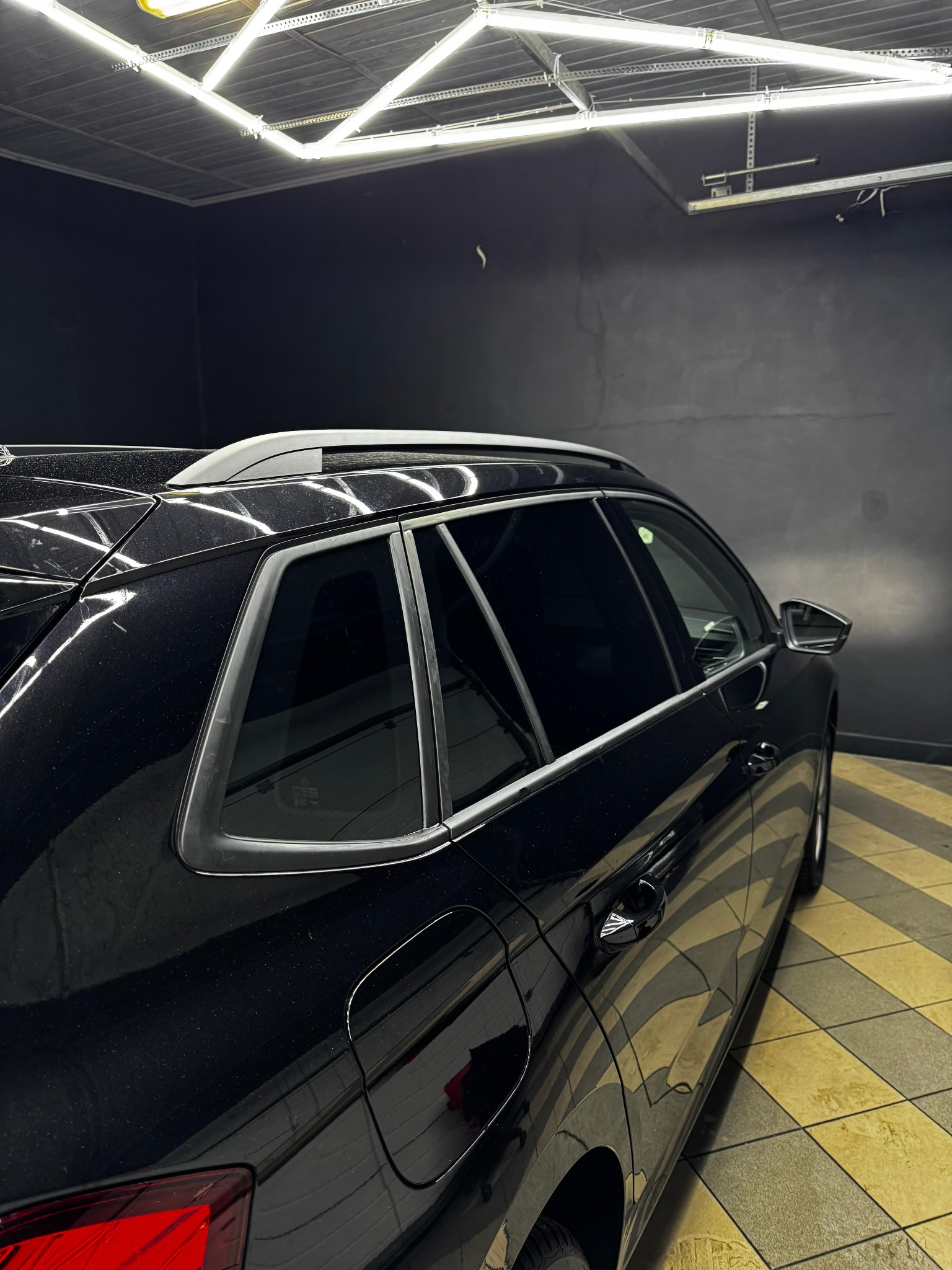 Częściowy widok czarnego samochodu typu SUV z przyciemnionymi szybami, zaparkowanego w dobrze oświetlonym garażu z wzorzystą podłogą.