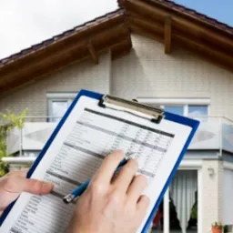 Osoba z niebieskim długopisem zaznacza pozycje w tabeli na niebieskim clipboardzie na tle jasnego domu z balkonem i anteną satelitarną.
