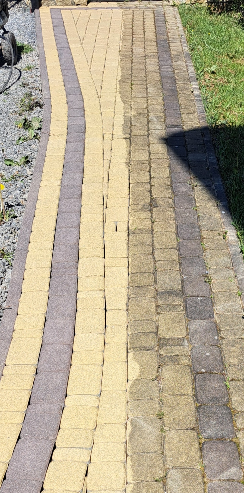 Dwukolorowa kostka brukowa, czyszczona połowicznie, z widoczną różnicą w odcieniu i zabrudzeniach między czystą a brudną częścią. Na obrzeżach widoczne kamienie i trawa.