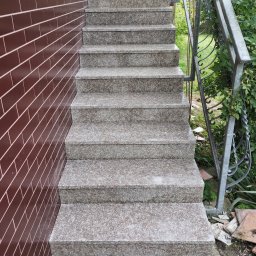 STYL-BUD KAZIMIERZ M - Granitowe schody zewnętrzne z metalową balustradą, przylegające do ściany wyłożonej brązowymi płytkami. Widoczne elementy zieleni w tle.
