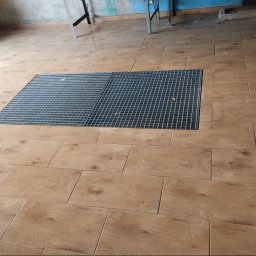 STYL-BUD KAZIMIERZ M - Ułożona podłoga z płytek imitujących drewno, z metalową kratką wentylacyjną wkomponowaną w posadzkę. Widoczne fragmenty rusztowania i ściany w tle.