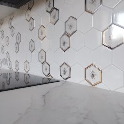 STYL-BUD KAZIMIERZ M - Białe, heksagonalne płytki z motywem pszczół, niektóre z obramowaniem w kolorze złota i srebra, tworzą unikalny backsplash w kuchni nad blatem z marmuru.