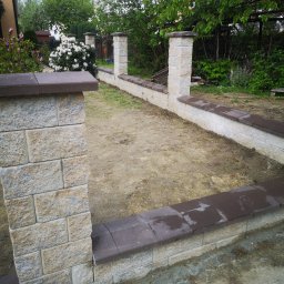 STYL-BUD KAZIMIERZ M - Nowo zbudowany murek ogrodzeniowy z beżowych bloczków kamiennych z ciemnymi, płaskimi nakrywami, otaczający pustą przestrzeń ziemi pod przyszły ogród. Widoczne trzy słupki z bloczków, w tle zielone...