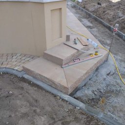 STYL-BUD KAZIMIERZ M - Budowa zewnętrznego podestu z beżowych płyt kamiennych, obok układana jasna kostka brukowa w łuk, widoczne narzędzia i niezakończone prace ziemne.