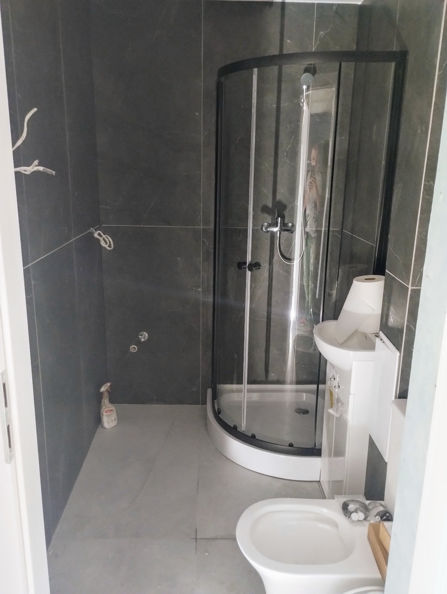 Szara łazienka z kabiną prysznicową z czarną ramą, umywalką, toaletą i butelką środka czyszczącego na podłodze; widoczne niedokończone podłączenia elektryczne na ścianie.