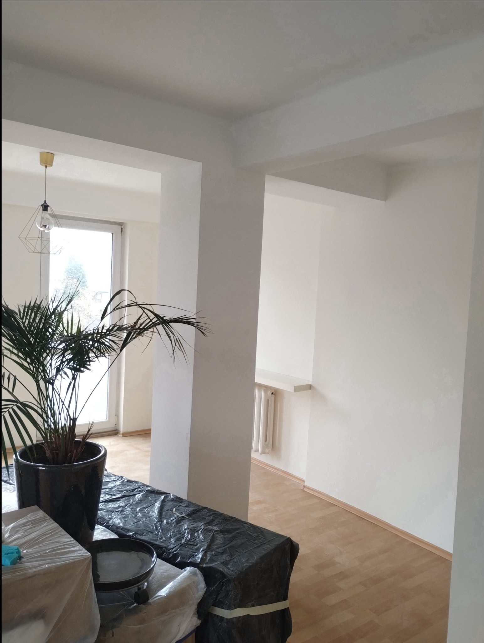 Przygotowania do malowania: Pokój z białymi ścianami, podłogą z paneli, zabezpieczonymi meblami folią, rośliną doniczkową i widocznymi śladami świeżo nałożonej gładzi na suficie.