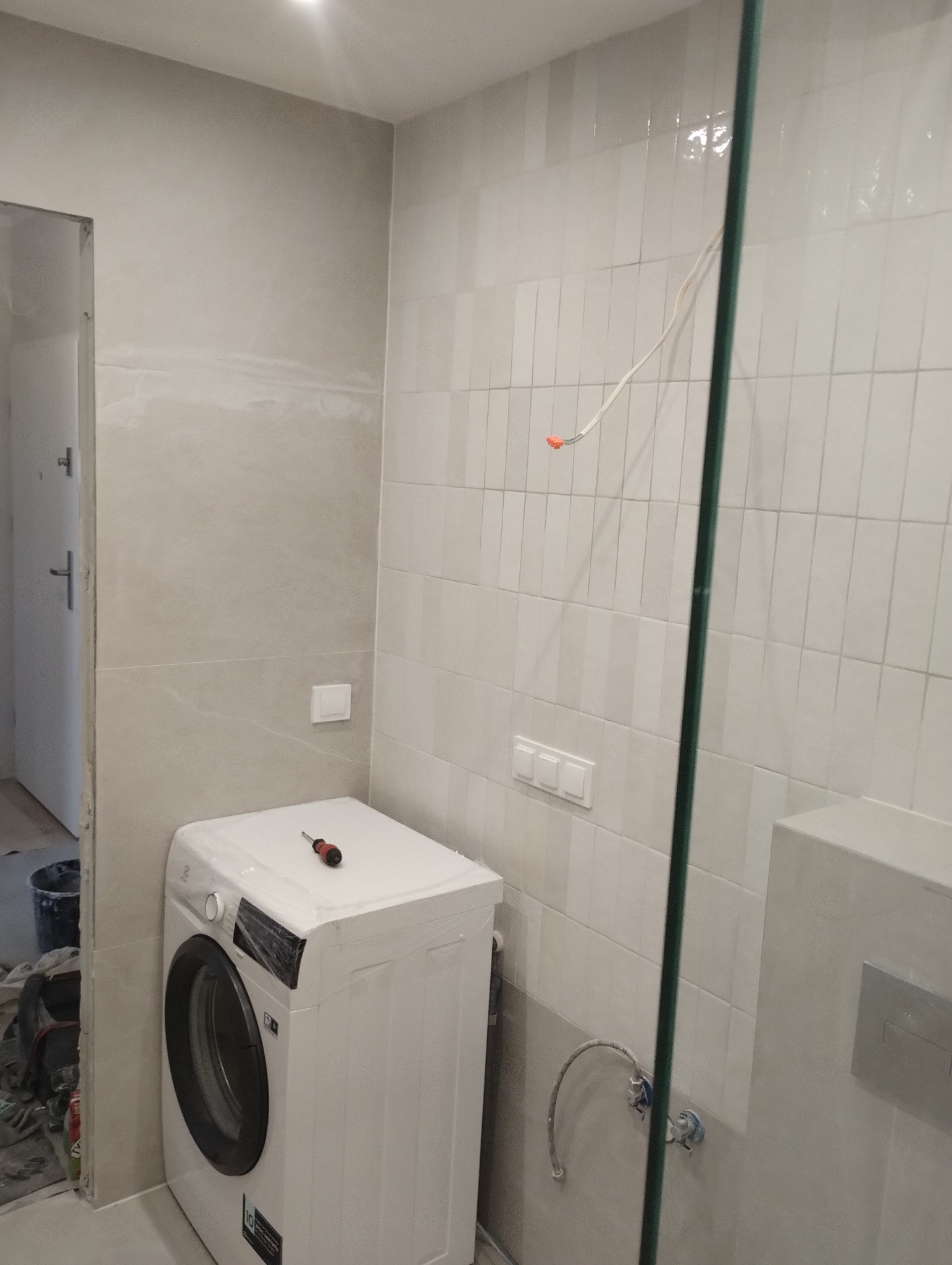 Wnętrze łazienki w trakcie remontu: widoczna pralka z narzędziem na górze, świeżo położone płytki w odcieniach szarości, wystające kable i fragment kabiny prysznicowej.