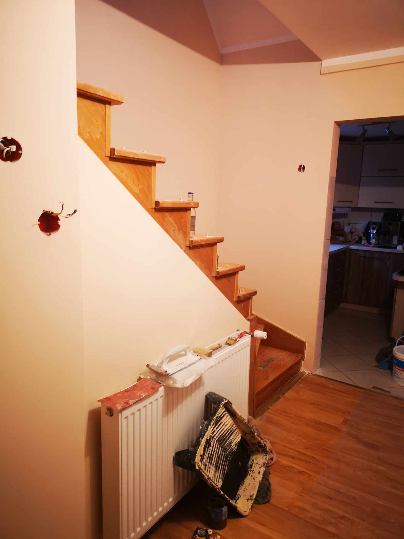 Ściana pomalowana na jasny beż z drewnianymi schodami w trakcie prac wykończeniowych, widoczne puszki elektryczne bez osprzętu i narzędzia malarskie przy grzejniku.