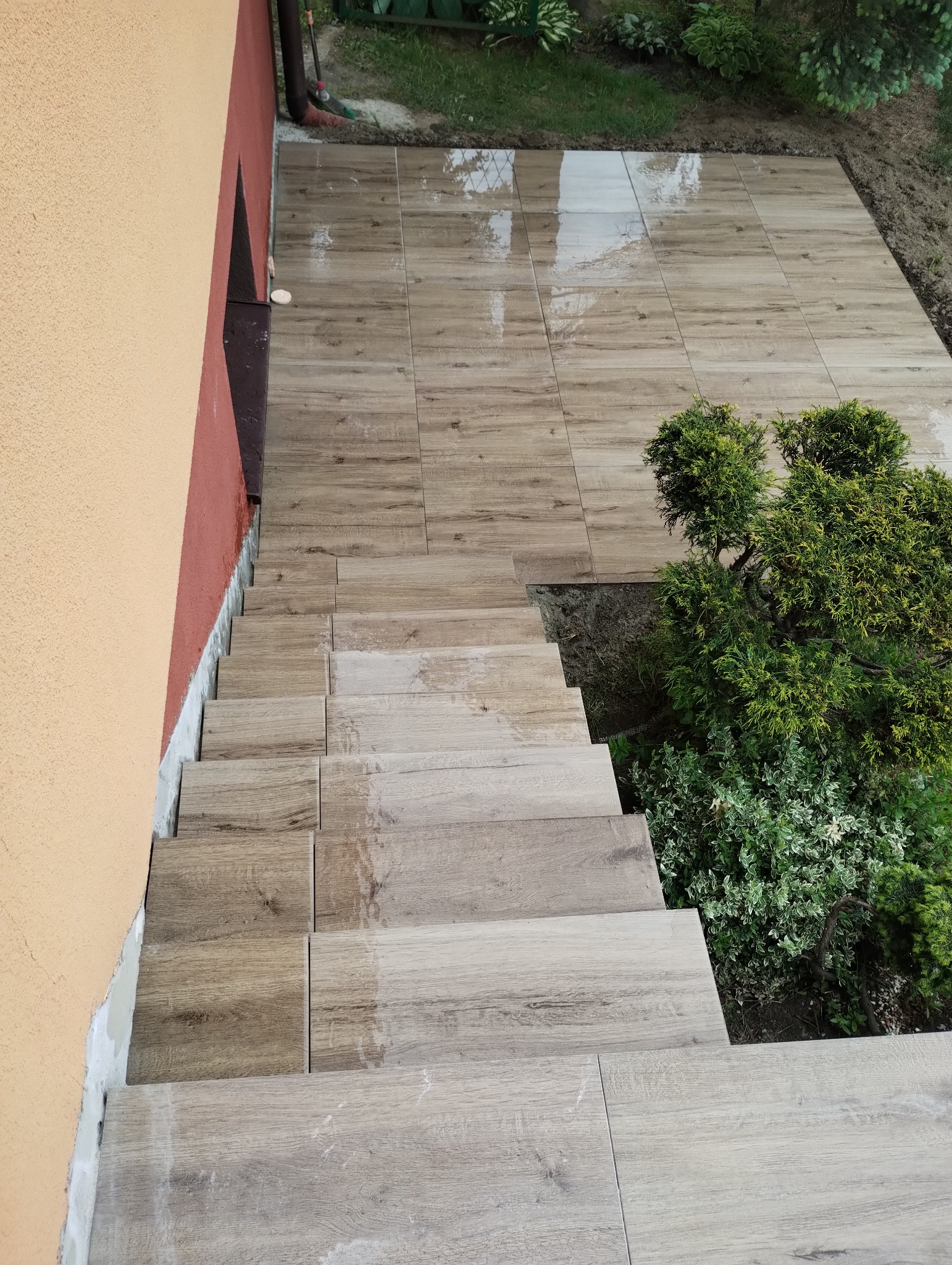Wykończone płytkami schody zewnętrzne z efektem drewna, prowadzące do ogrodu. Widok z góry, z fragmentem elewacji budynku w tle. Płytki ułożone schludnie, z zielenią po prawej stronie.