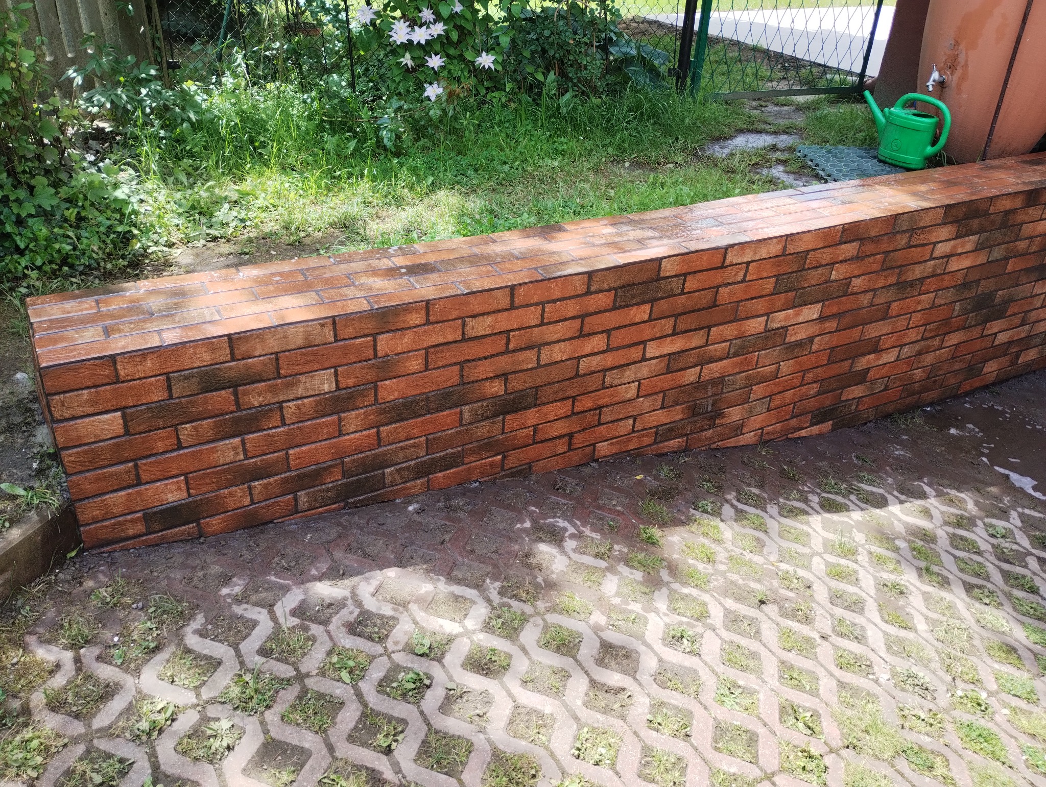 Ściana z płytek imitujących cegłę w ogrodzie, widoczna zieleń i konewka. Wykończenie zewnętrzne z użyciem płytek, imitacja cegły, dekoracja ogrodu.