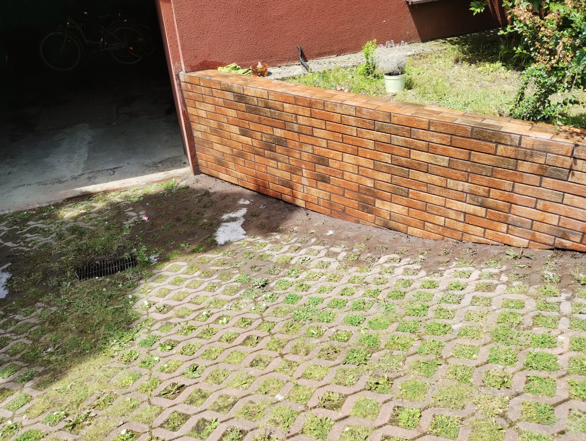 Ściana z cegły klinkierowej obok garażu, z roślinnością na górze. Kostka ażurowa na ziemi. Widoczne rowy odpływowe.