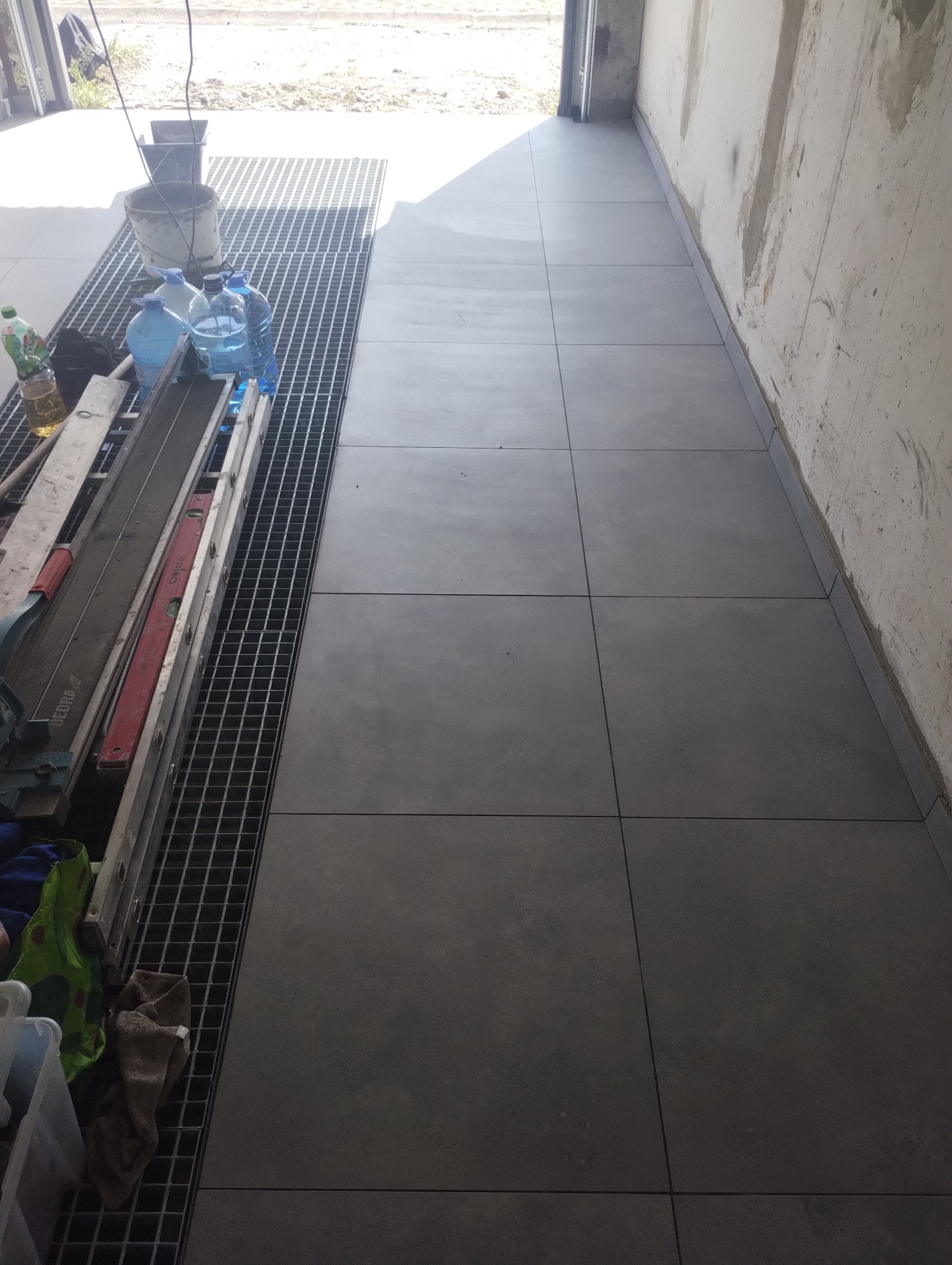 Ułożone, szare płytki podłogowe w korytarzu, widoczne narzędzia pracy i materiały budowlane. Minimalistyczne wykończenie, surowe ściany w tle.