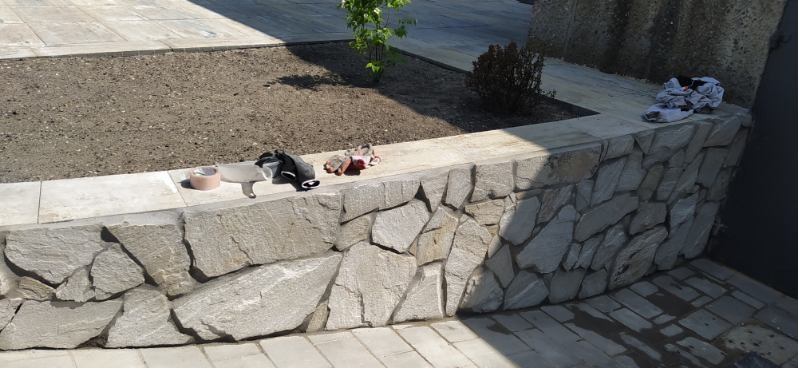 Kamienna ścianka rabaty kwiatowej z narzędziami, świeżo po ułożeniu, na tle kostki brukowej i zieleni. Detal wykończenia ogrodu z użyciem naturalnego kamienia.