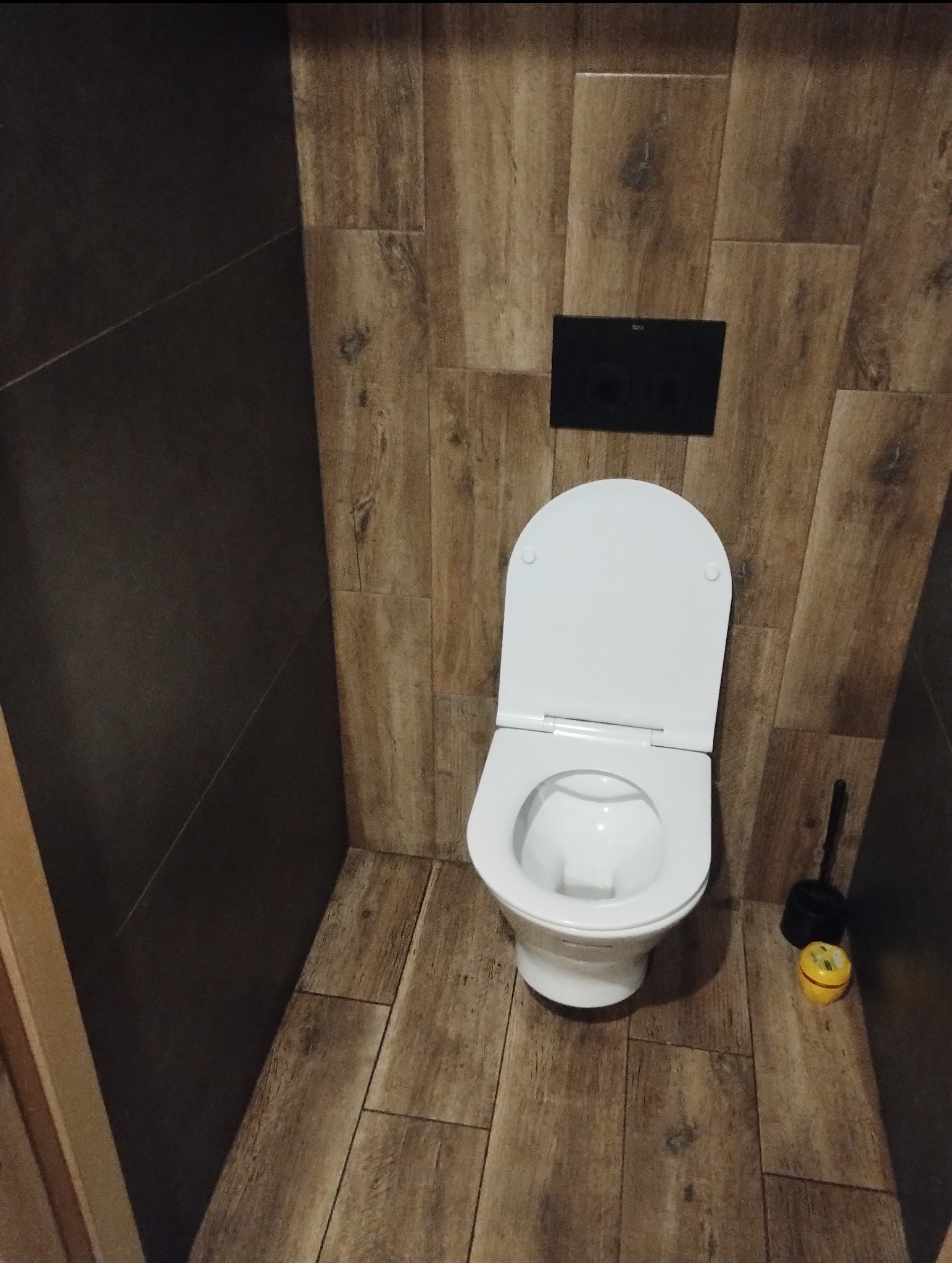 Nowoczesna toaleta z deską sedesową w jasnej łazience wykończonej płytkami imitującymi drewno, z czarnym przyciskiem spłukującym i akcesoriami.