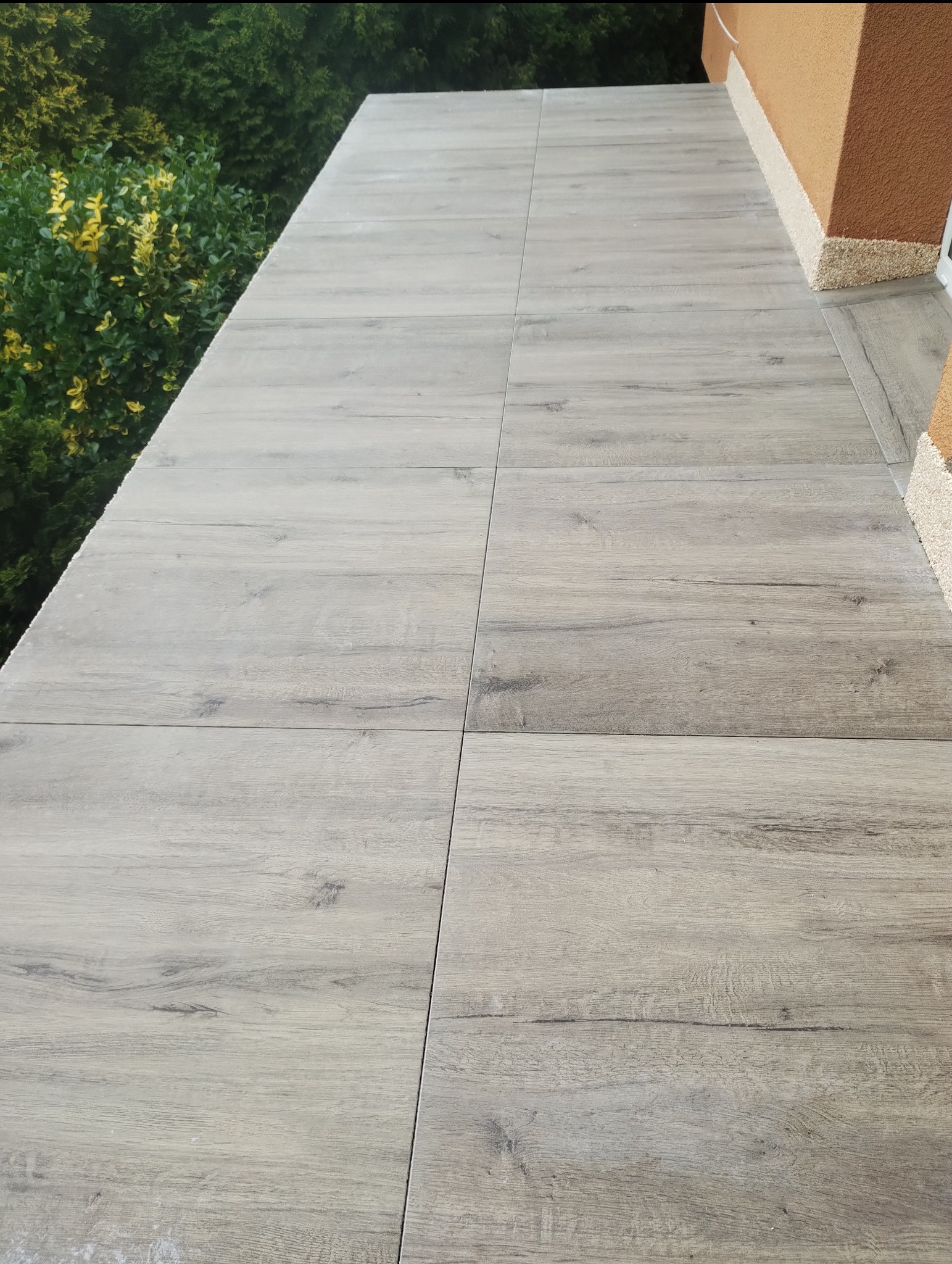 Jasne, drewnopodobne płytki gresowe ułożone na balkonie, widok z góry. Zielone krzewy w tle, ściana budynku z beżowym wykończeniem.