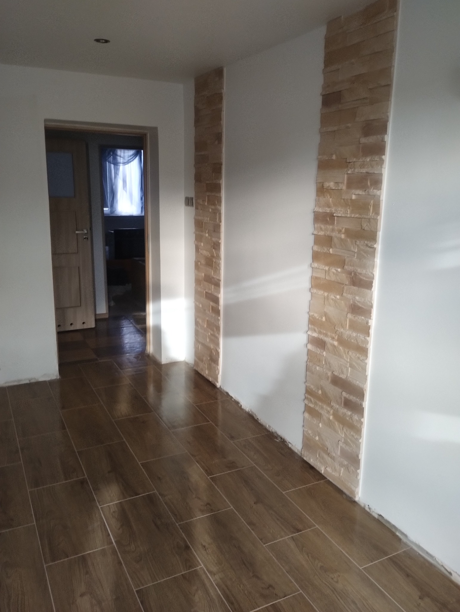 Ukończone wnętrze: brązowe płytki podłogowe imitujące drewno, fragment ściany z dekoracyjnym kamieniem w odcieniach beżu, widok na drzwi i okno w tle.