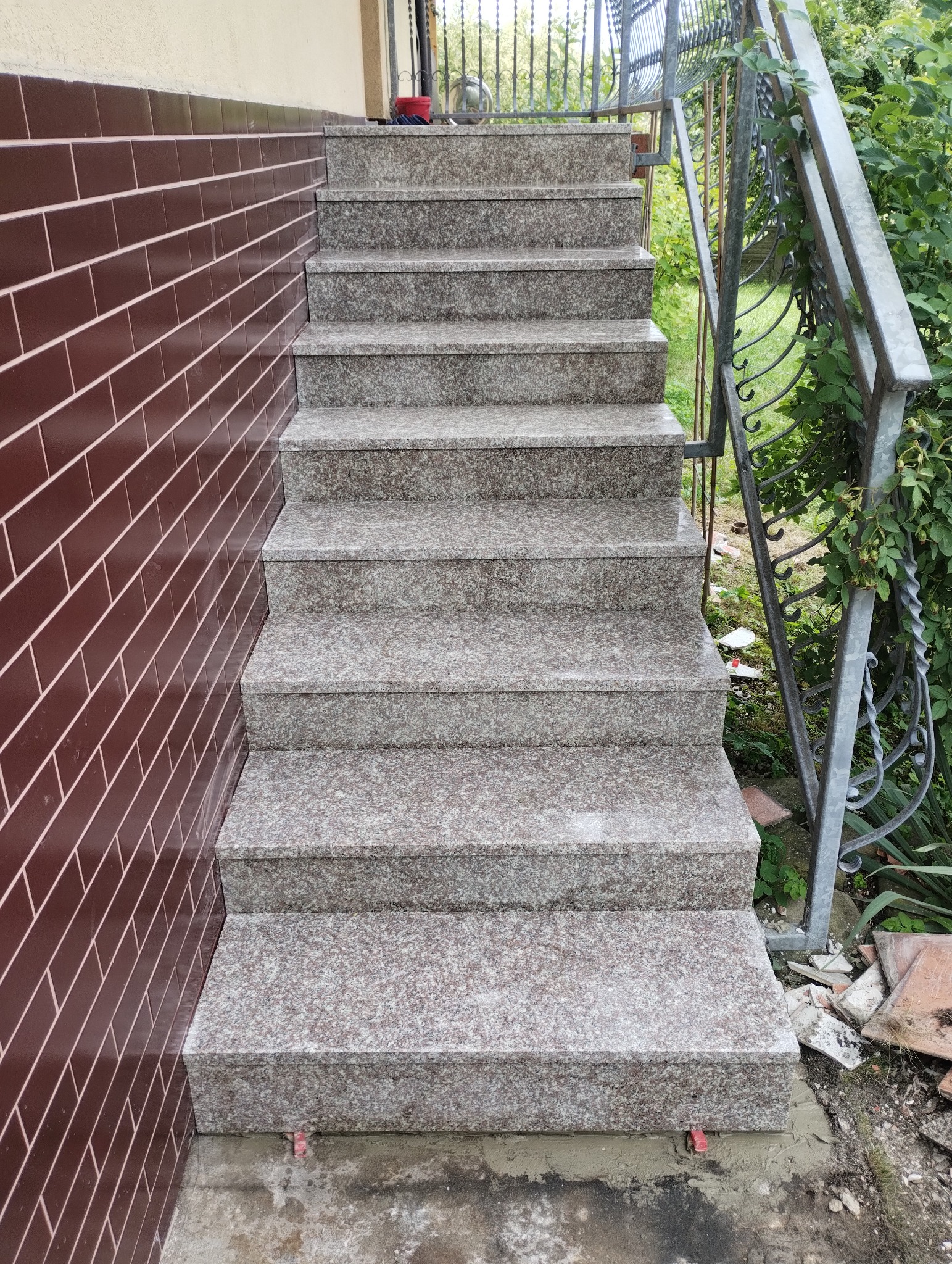 Granitowe schody zewnętrzne z metalową balustradą, przylegające do ściany wyłożonej brązowymi płytkami. Widoczne elementy zieleni w tle.
