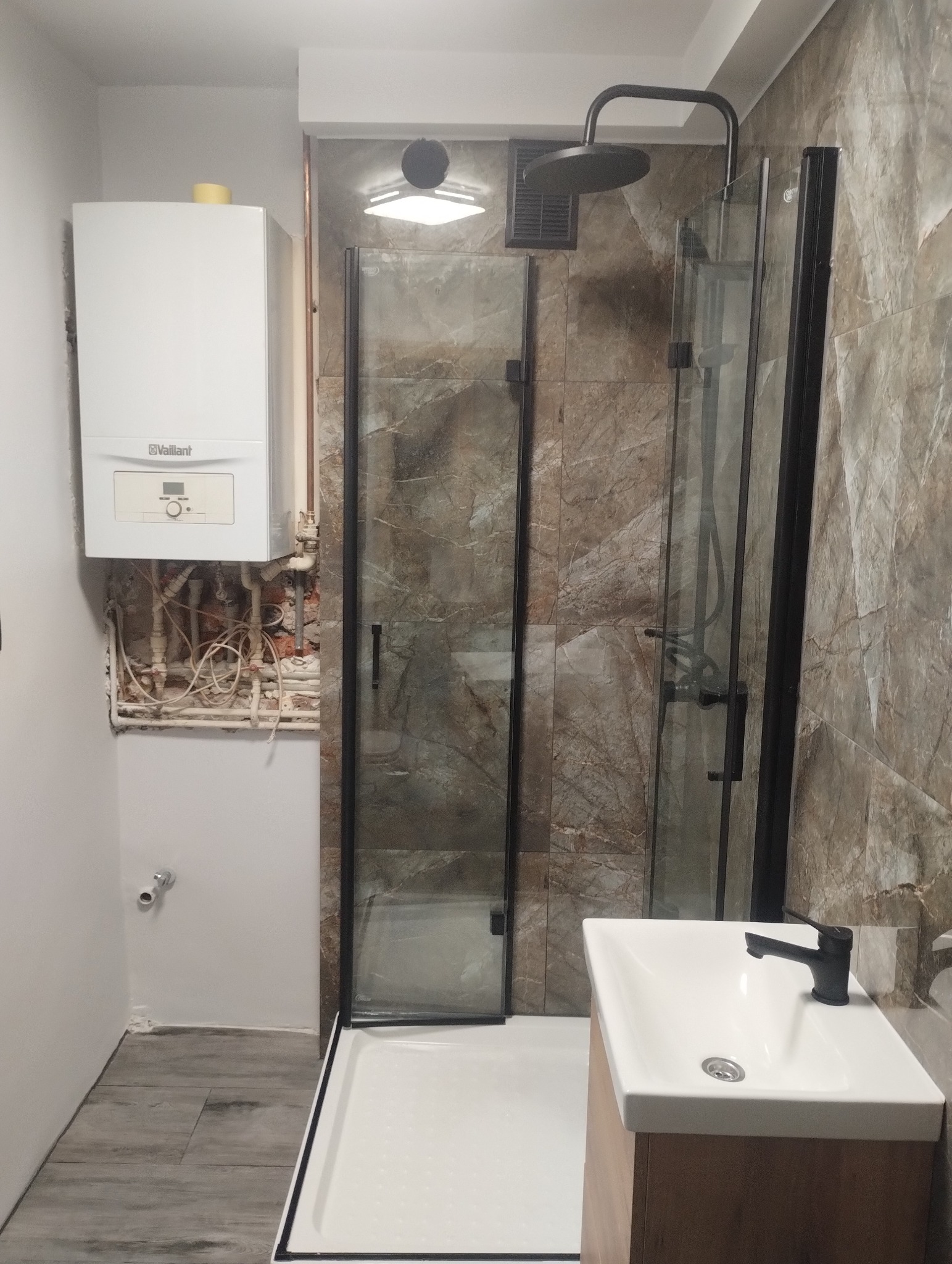 Nowoczesna kabina prysznicowa z czarnymi profilami i deszczownicą, umywalka z szafką, piec gazowy Vaillant na ścianie. Widoczne niedokończone prace instalacyjne.