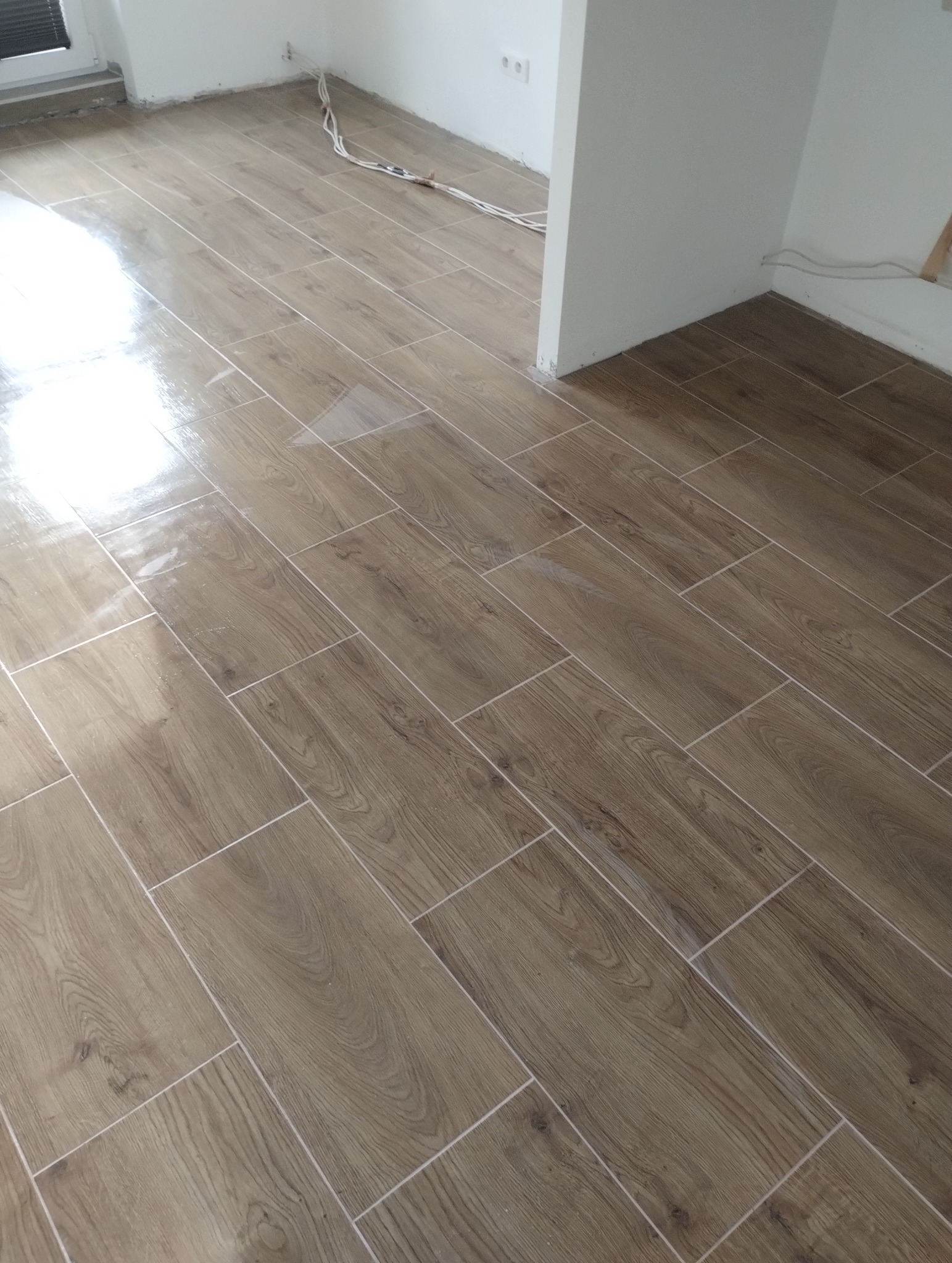 Ułożona podłoga z płytek imitujących drewno w jasnym pomieszczeniu. Widoczne kable elektryczne przy ścianie. Nowoczesny wzór, świeżo po remoncie.