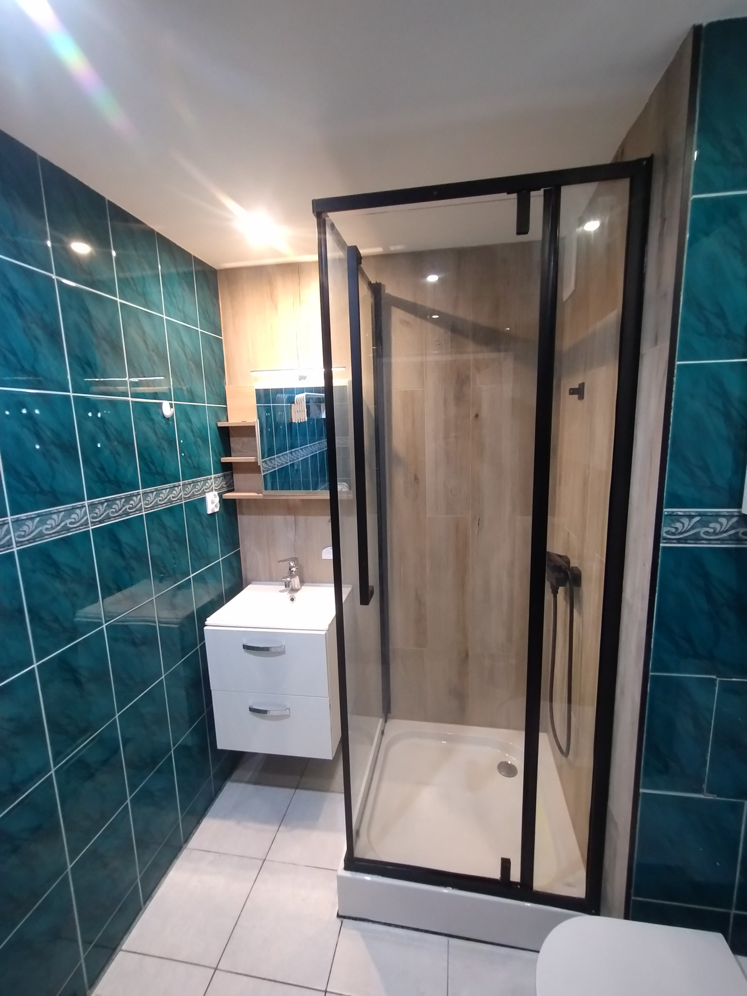 Wykończona łazienka z kabiną prysznicową w czarnej ramie, umywalką z szafką i turkusowymi płytkami. Widoczne detale wykończenia i oświetlenie.