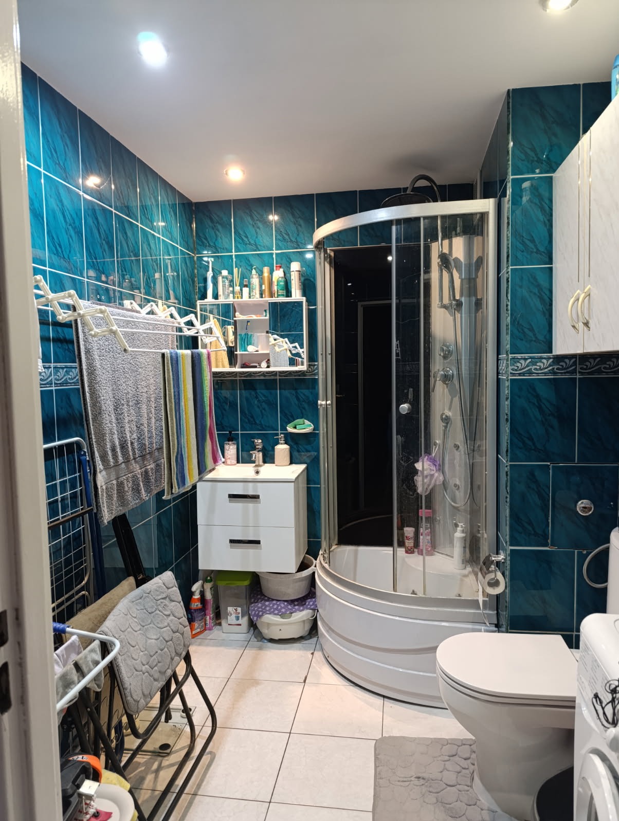 Niebieska łazienka z kabiną prysznicową, białą szafką pod umywalką i wiszącą suszarką na ręczniki. Widoczne akcesoria łazienkowe i płytki w odcieniach turkusu.