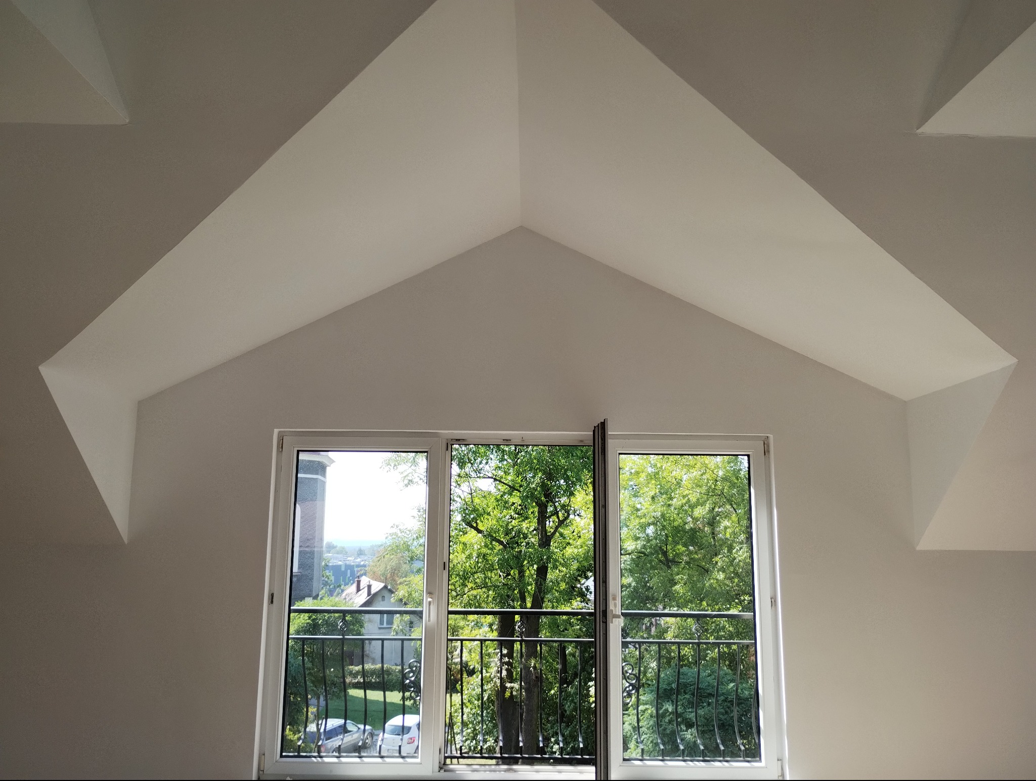 Poddasze z oknem balkonowym i widokiem na zieleń. Ściany i sufit w kolorze białym, świeżo po malowaniu. Charakterystyczna geometria skosów.