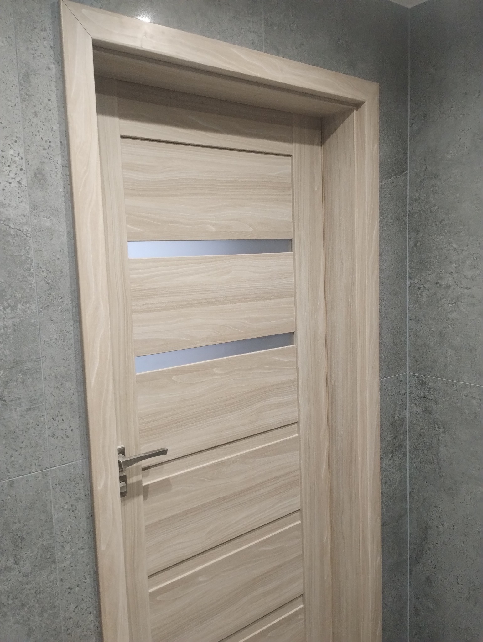 Zamknięte, jasne drzwi wewnętrzne z poziomymi przeszkleniami, zamontowane w łazience z szarymi płytkami. Widoczna klamka i fragment ściany.