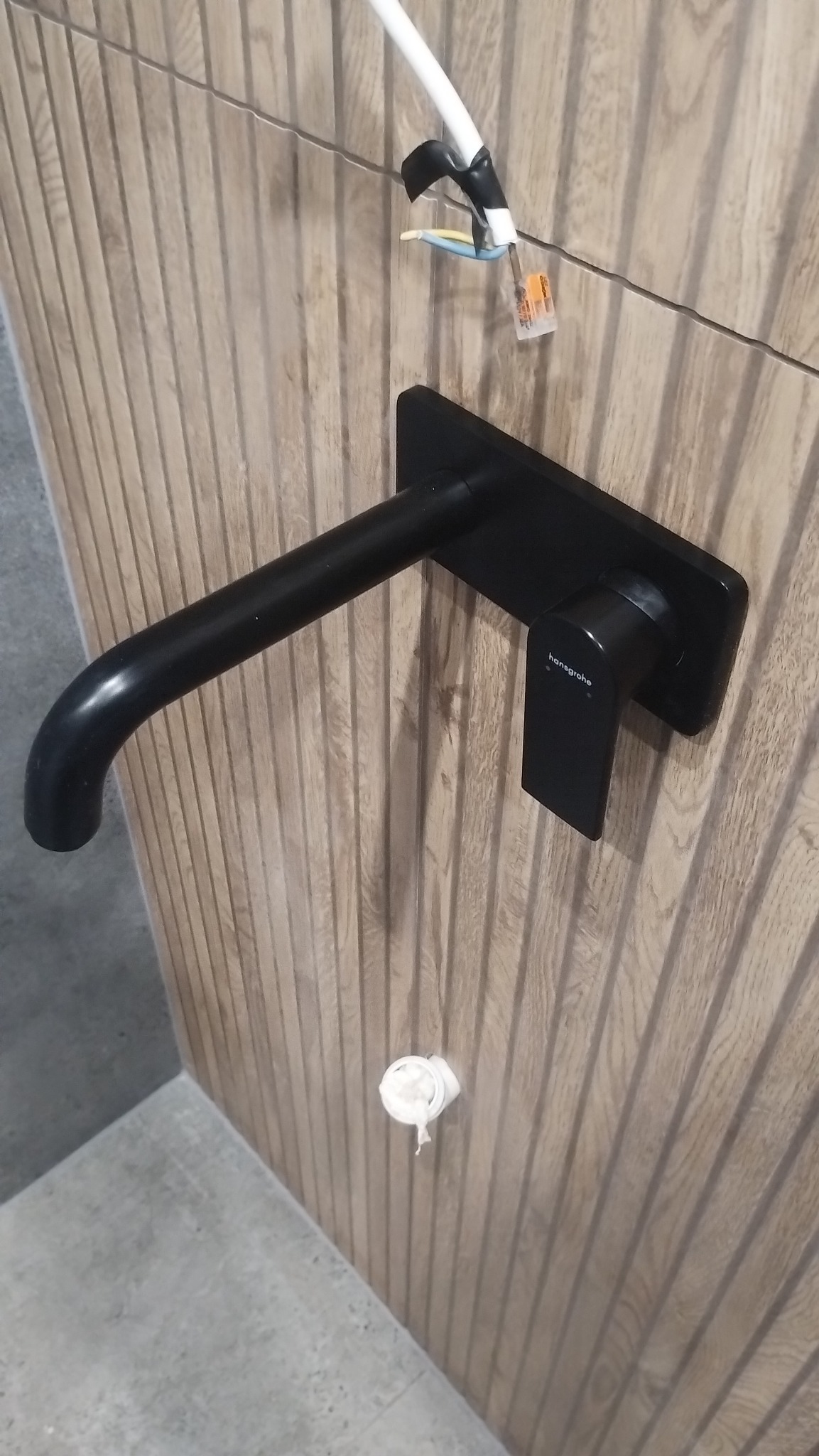 Czarna bateria Hansgrohe zamontowana na ścianie z drewnopodobnych paneli w łazience. Widoczne przyłącza elektryczne i fragment podłogi. Ujęcie z dołu.