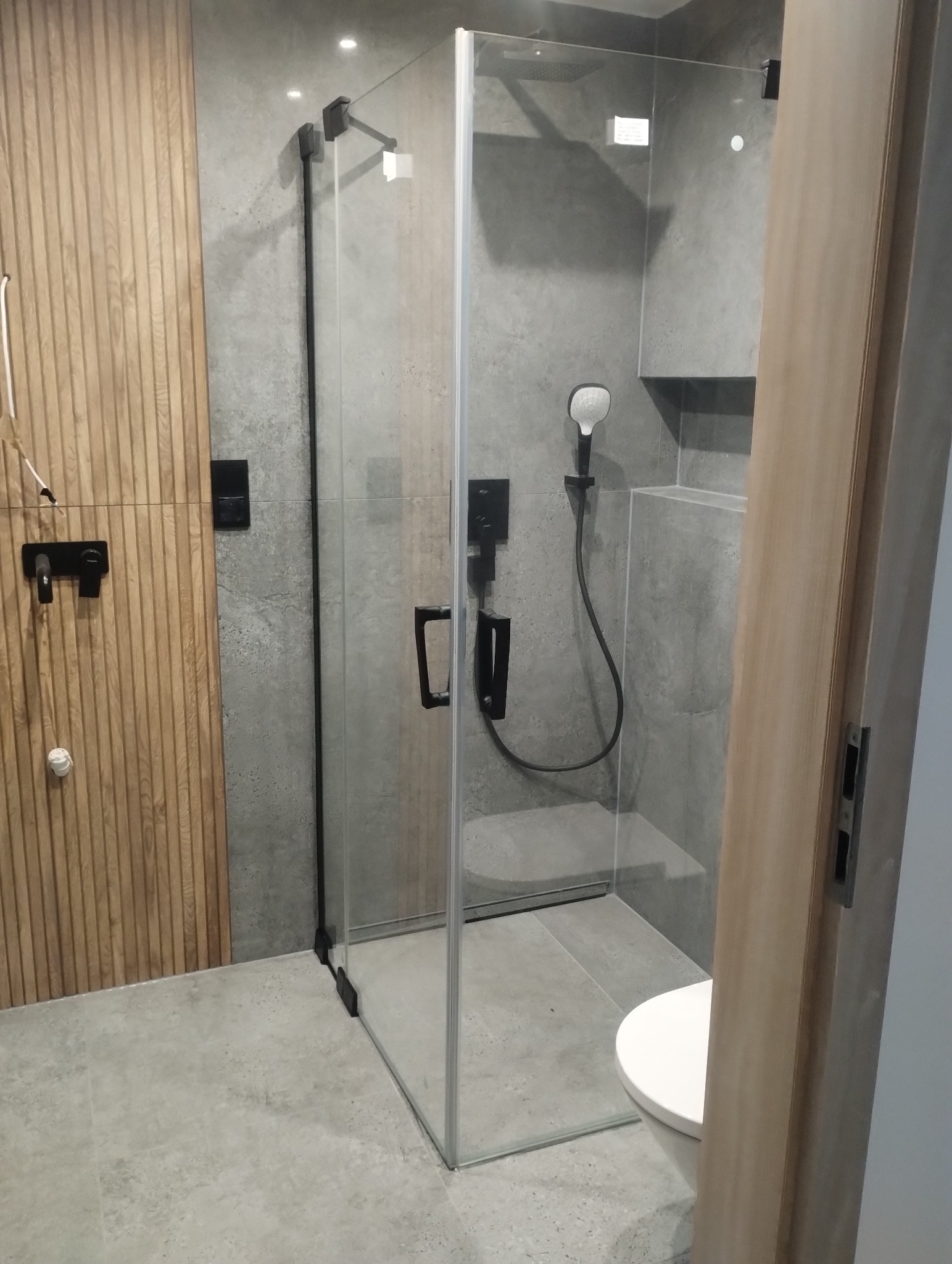 Nowoczesna kabina prysznicowa z czarnymi elementami armatury i drewnianą ścianą w wykończonej łazience. Minimalistyczny design, szare płytki, częściowo widoczna toaleta.