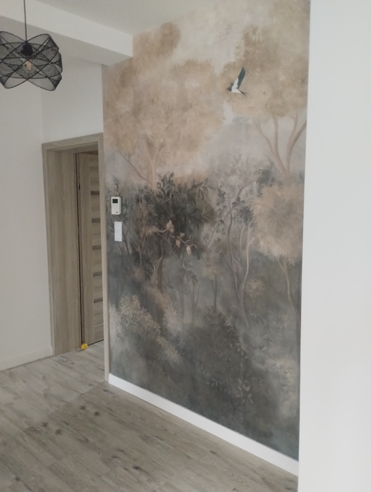 Ściana z malowidłem leśnym i ptakiem w nowocześnie wykończonym wnętrzu. Widoczny fragment podłogi i drzwi. Dekoracyjne oświetlenie sufitowe.