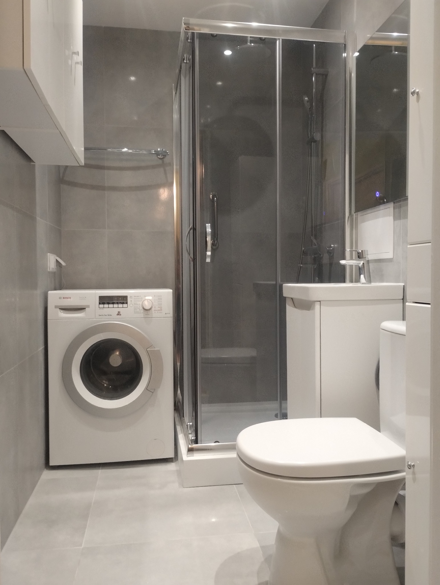 Nowoczesna łazienka w szarościach: kabina prysznicowa, umywalka z szafką, toaleta i pralka Bosch. Minimalistyczny design, płytki podłogowe i ścienne w odcieniach szarości.
