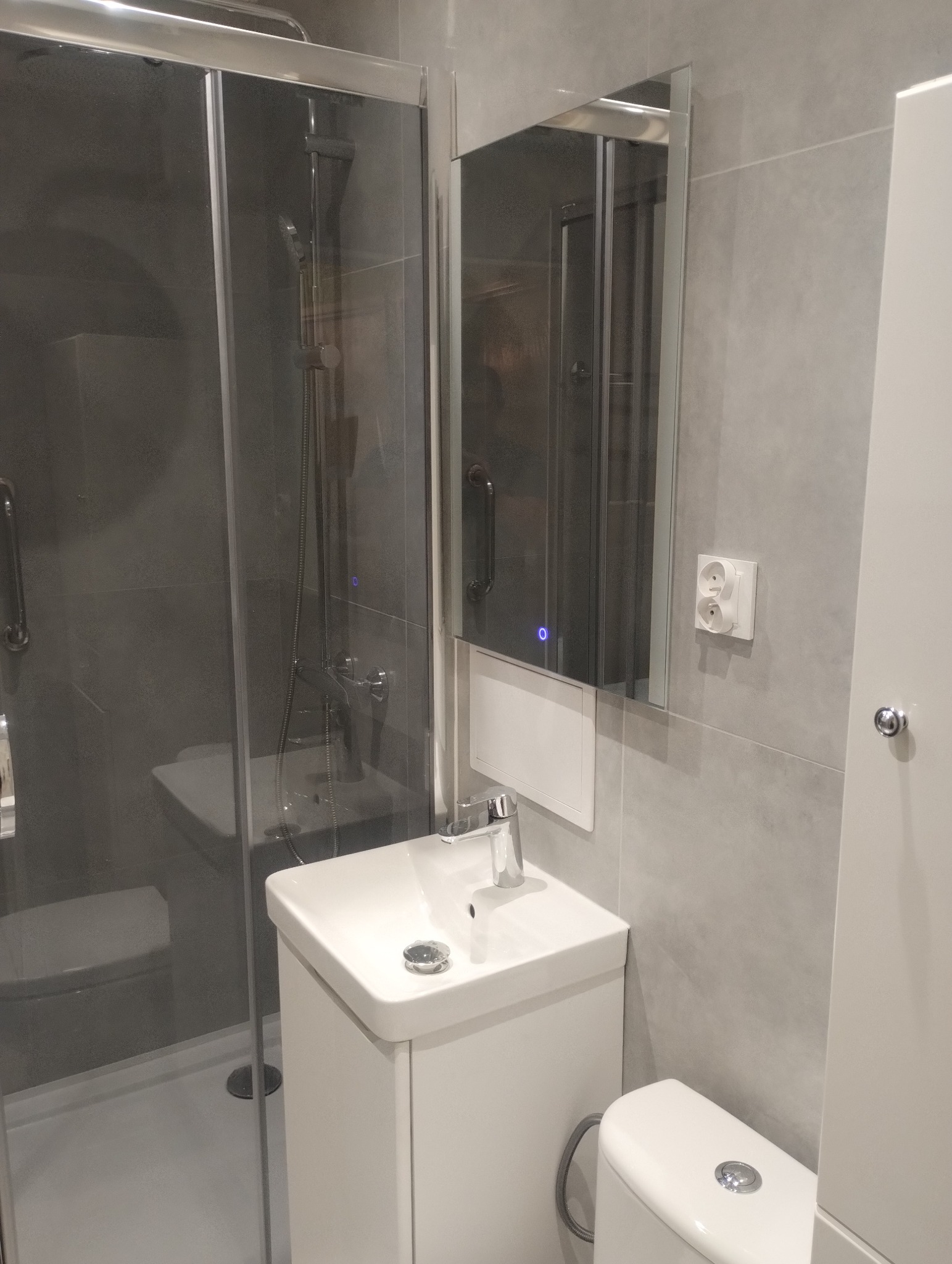 Nowoczesna łazienka w szarościach: kabina prysznicowa ze srebrnymi profilami, biała umywalka z szafką i lustro z podświetleniem LED. Minimalistyczny design.