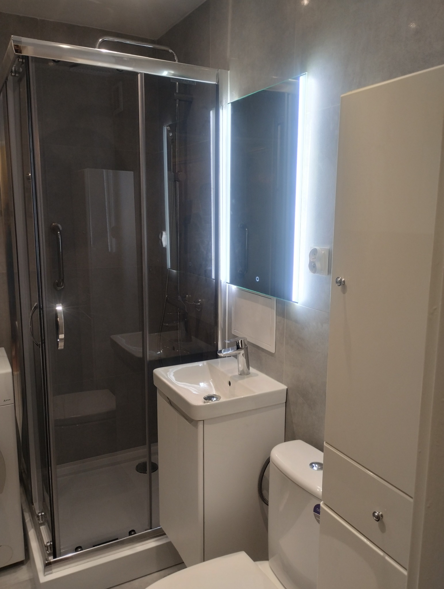 Nowoczesna łazienka z kabiną prysznicową, umywalką z szafką, toaletą i podświetlanym lustrem. Szare płytki na ścianach. Minimalistyczny design.