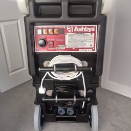 Łowcy Porządku - Firma Sprzątająca - Profesjonalny odkurzacz piorący Ashby's Enforcer V2 SteamMate z widocznym panelem sterowania, zwiniętym kablem zasilającym i wężami, stojący na szarym dywanie.