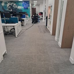 Łowcy Porządku - Firma Sprzątająca - Szeroki kadr na biuro typu open space z szarą wykładziną dywanową, na której widoczny jest sprzęt do czyszczenia dywanów z długim kablem, biurka z komputerami w tle i dekoracja ścienna z motywem...