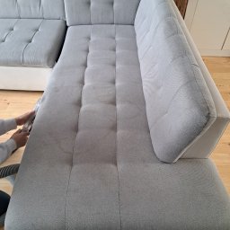 Łowcy Porządku - Firma Sprzątająca - Czyszczenie jasnoszarej, narożnej kanapy z tapicerką w delikatny wzór, widoczne ręce osoby czyszczącej oraz końcówka odkurzacza piorącego.