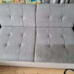 Łowcy Porządku - Firma Sprzątająca - Fragment szarej, tapicerowanej kanapy z widoczną różnicą w czystości po praniu na dwóch segmentach siedziska.